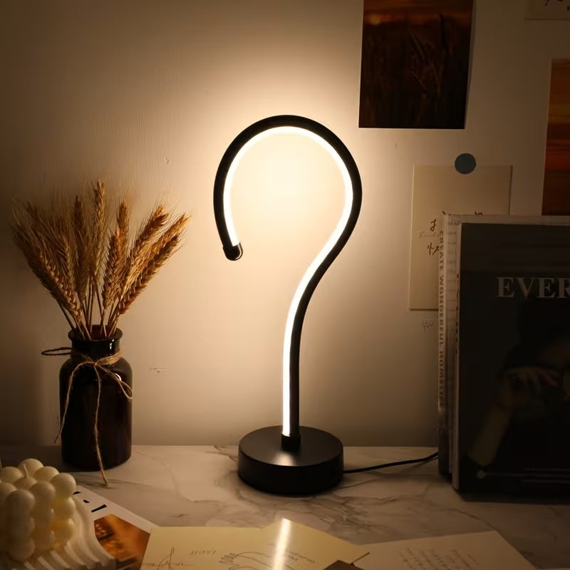 Modern table lamp with curved design on a desk next to a vase of wheat and books.